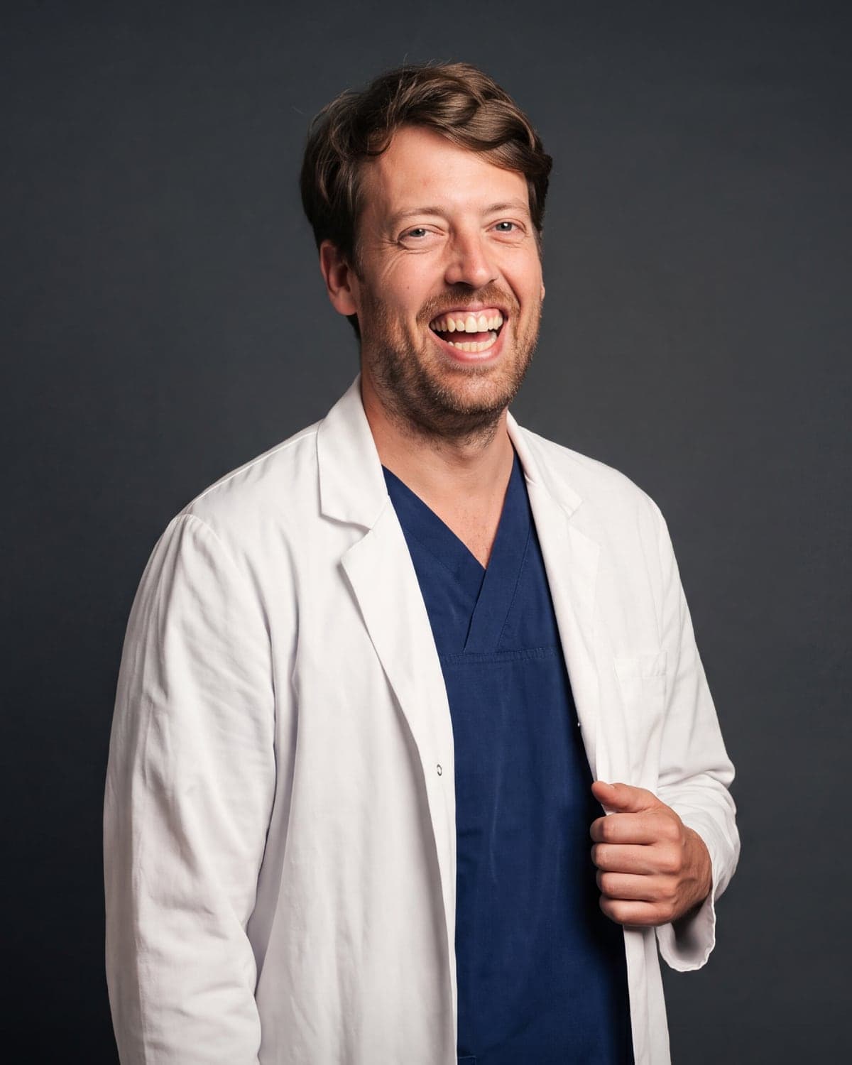 Dr. Elio Díez-Feijóo, medical director of AdmiraVisión and adéu.now, laughing in a white coat and navy scrubs against a dark studio backdrop