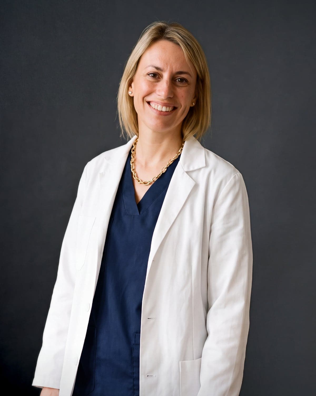 Dr. Pilar Prats, refractive surgeon at adéu.now, smiling in a white coat and navy scrubs against a dark studio backdrop