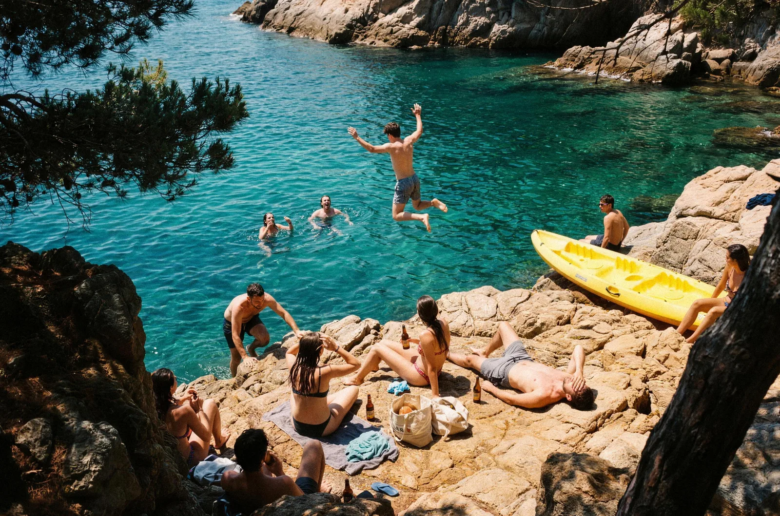 Amics en una cala de roques de la costa catalana: un salta al mar turquesa, dos neden i la resta pren el sol a les roques